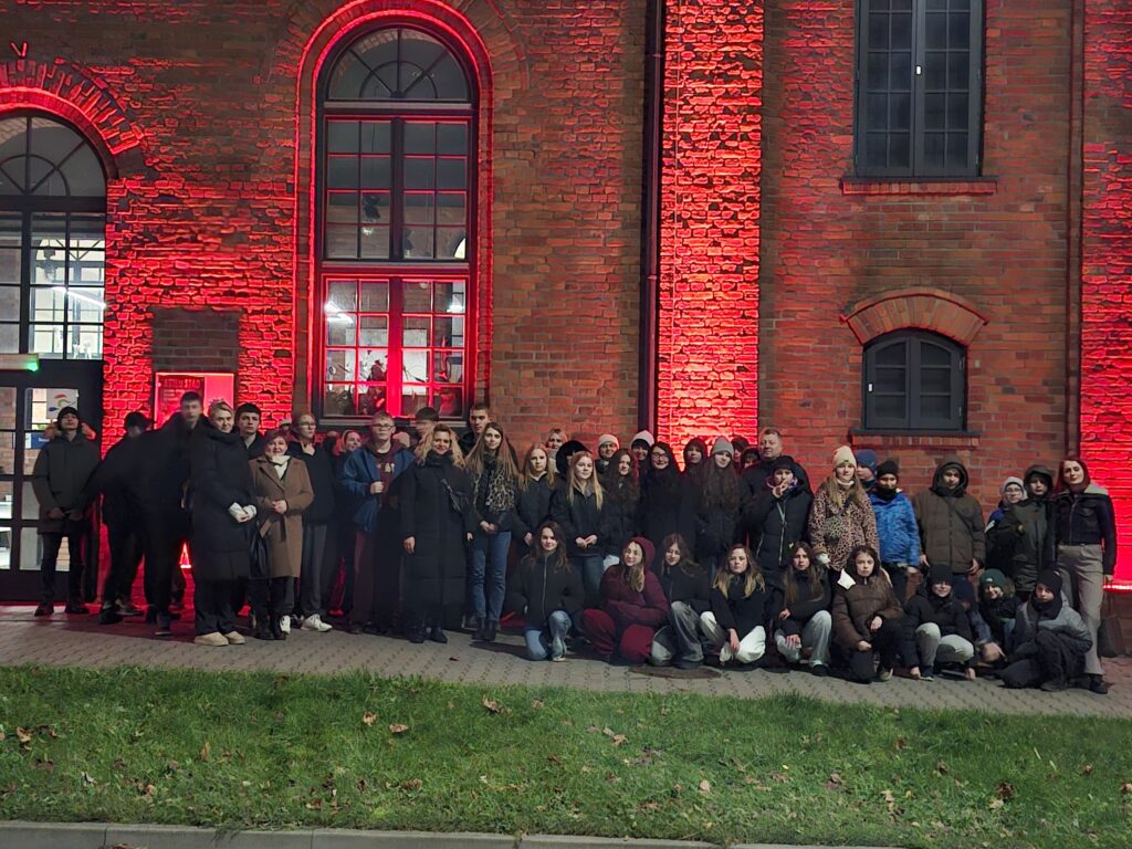 Podświetlony na czerwono budynek browaru przed którym stoi do zdjęcia duża grupa ludzi.