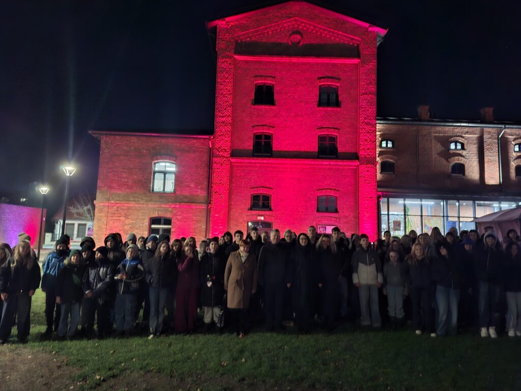Podświetlony na czerwono budynek browaru przed którym stoi do zdjęcia duża grupa ludzi.