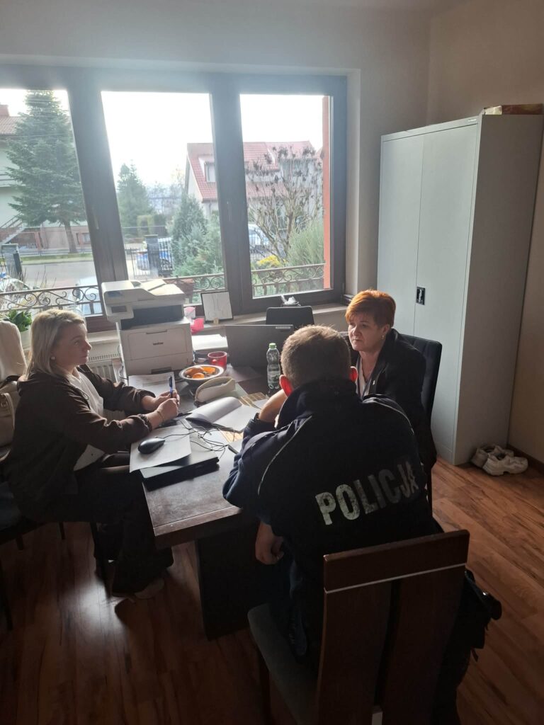 Dwie osoby z policjantem rozmawiają przy stole na tle oświetlonego okna.